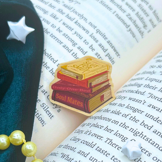 A small enamel pin shaped like a stack of three books with the words 'Soul Mates' and 'Only One Bed' on the covers, placed on open book pages next to a dark navy star-shaped accessory and pale yellow pearls.