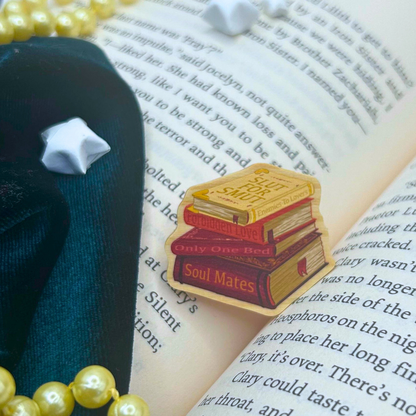 A small enamel pin shaped like three stacked books with the words 'Soul Mates' and 'Only One Bed' on the spines, placed on an open book with decorative props including pearl beads and a dark fabric.