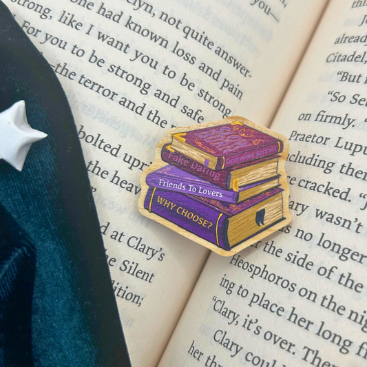 A die-cut enamel pin shaped like a stack of four books with purple and gold covers, featuring the words 'Fake Lovers', 'Friends To Lovers', and 'WHY CHOOSE?' printed on the spines. The pin is styled on an open book with a dark navy blue sleeve visible on the left side.