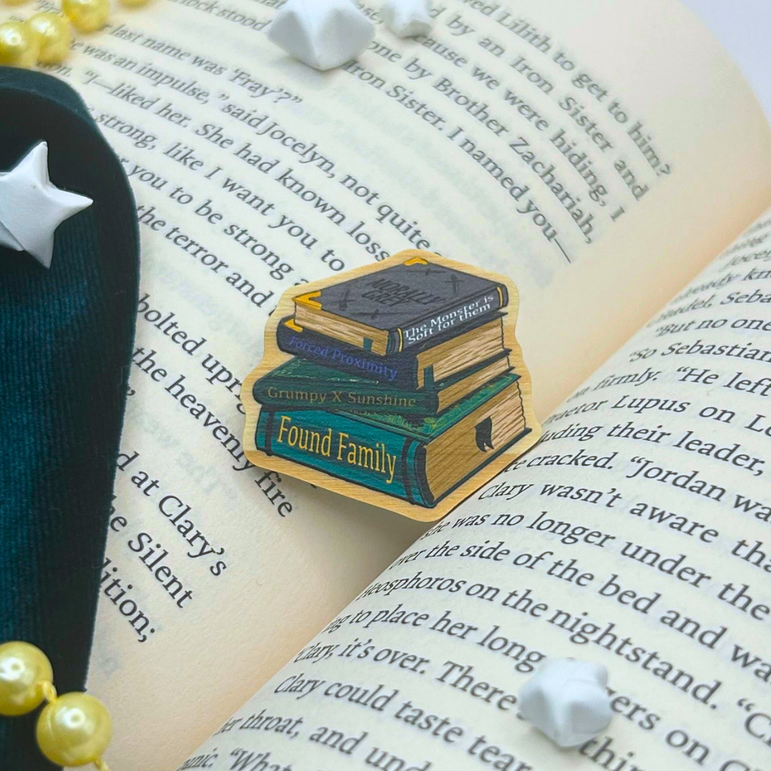 A die-cut enamel pin shaped like a stack of three books with the words 'Found Family' on the front, displayed on an open book with pearl beads and white stones around it.