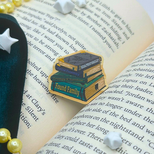 A die-cut enamel pin shaped like a stack of three books with the words 'Found Family' on the front, displayed on an open book with pearl beads and white stones around it.