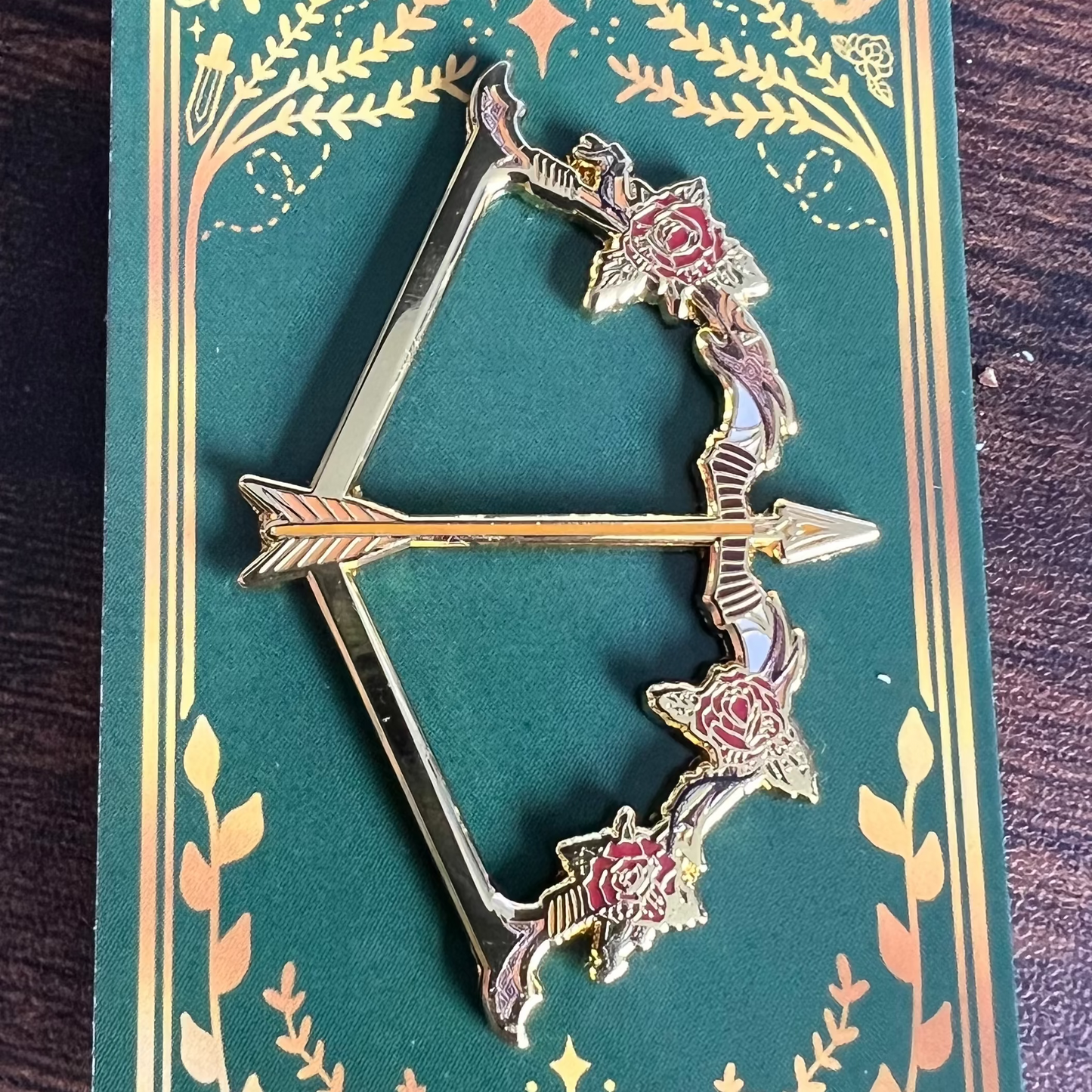 Decorative bow and arrow pin on a green card with floral designs, on a wooden surface.